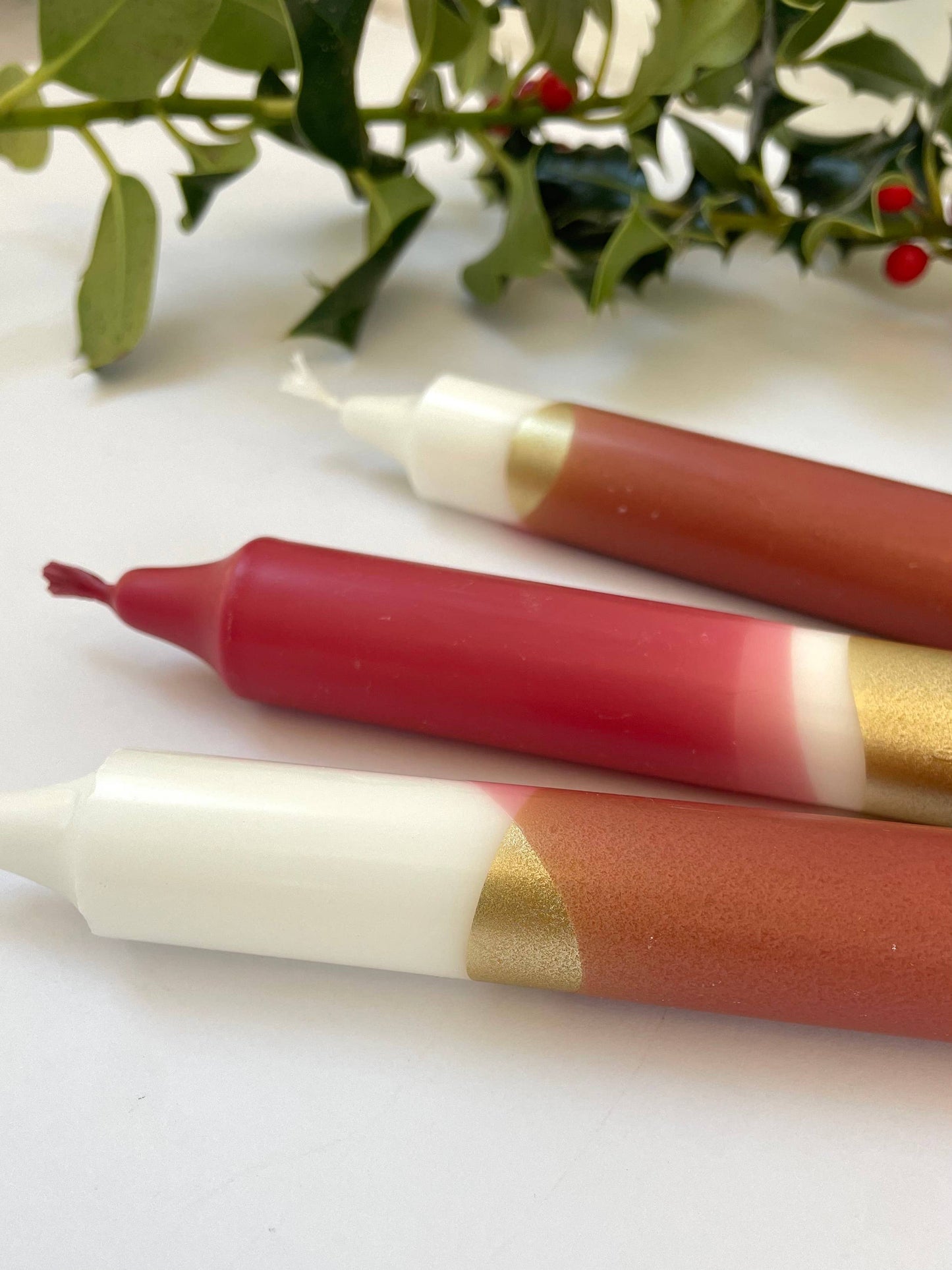 Three candles with red, white, and gold bands on a white surface with green foliage in the background by The Singing Rabbit for Heart Gallery
