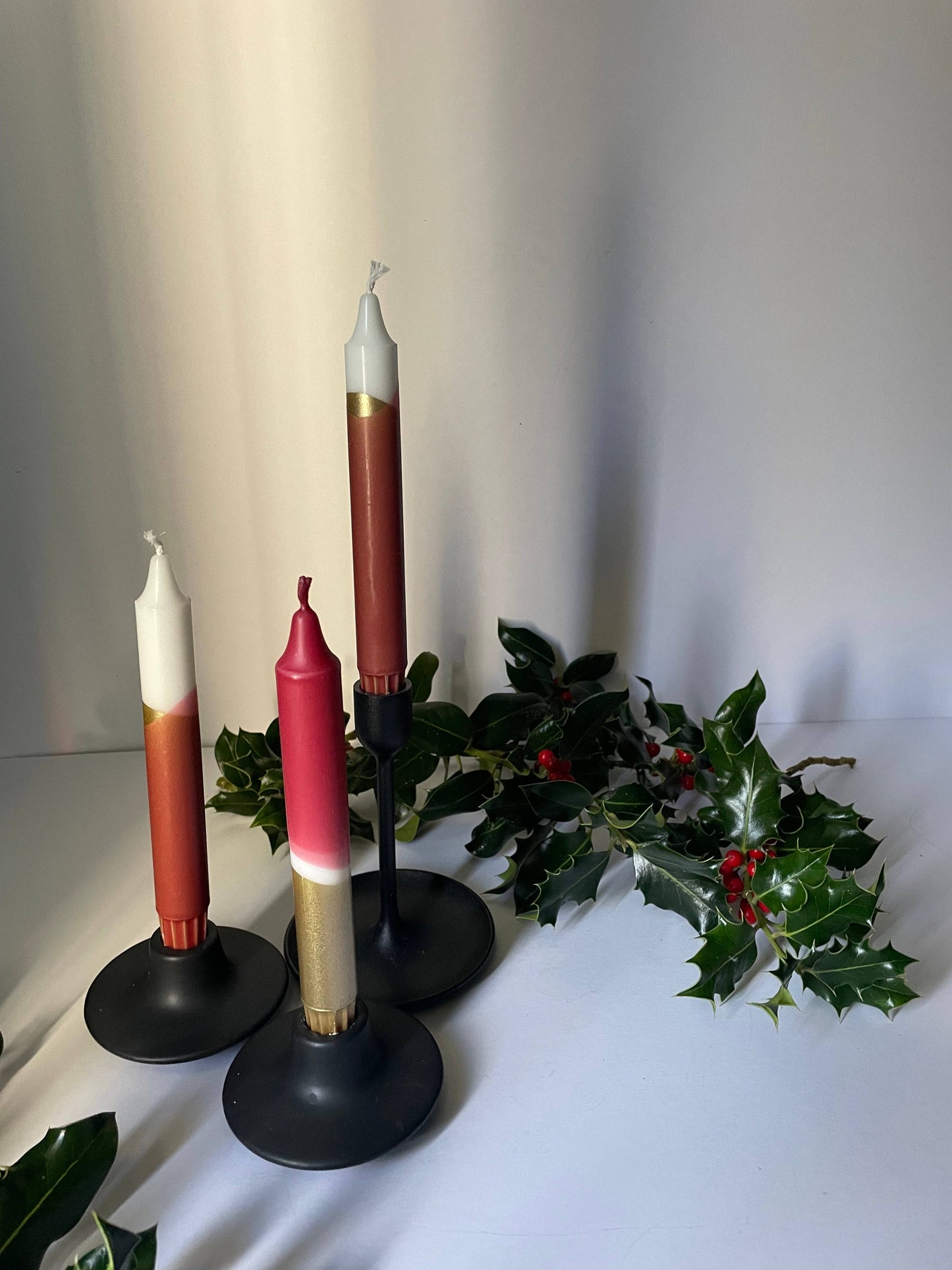 Three candlesticks with different colored candles on a white surface with greenery.