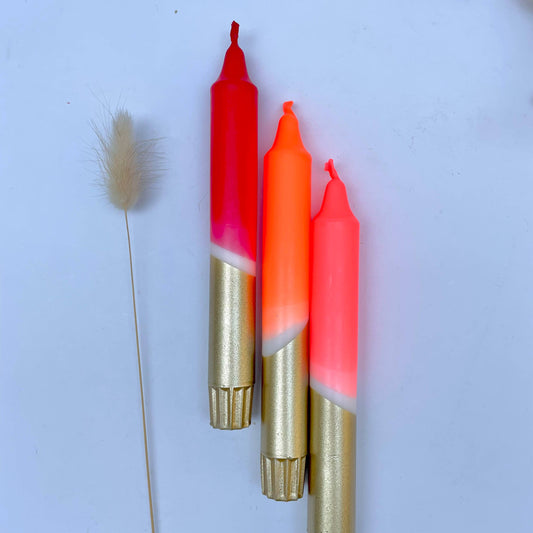 Three dip-dyed dinner candles with gold-colored bases and neon pink to orange gradients, displayed on a white surface alongside a dry plant stem.
