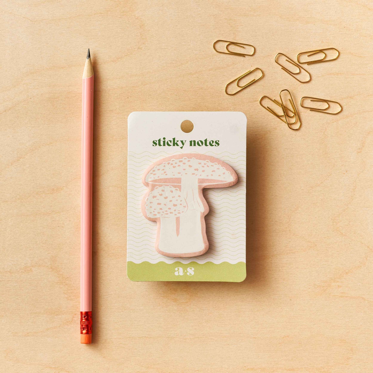 A pack of mushroom-shaped sticky notes by Another Studio with a single pink pen and multiple gold paper clips on a wooden surface.