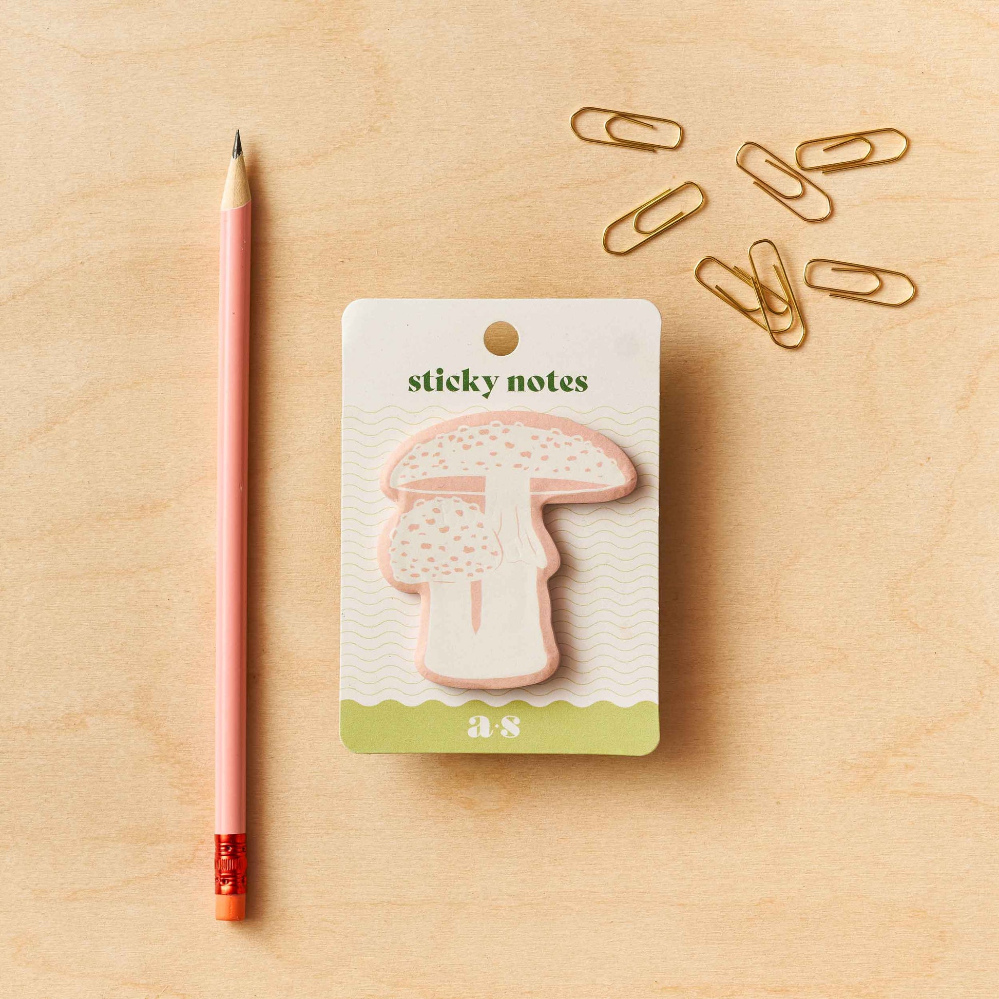 A pack of mushroom-shaped sticky notes by Another Studio with a single pink pen and multiple gold paper clips on a wooden surface.