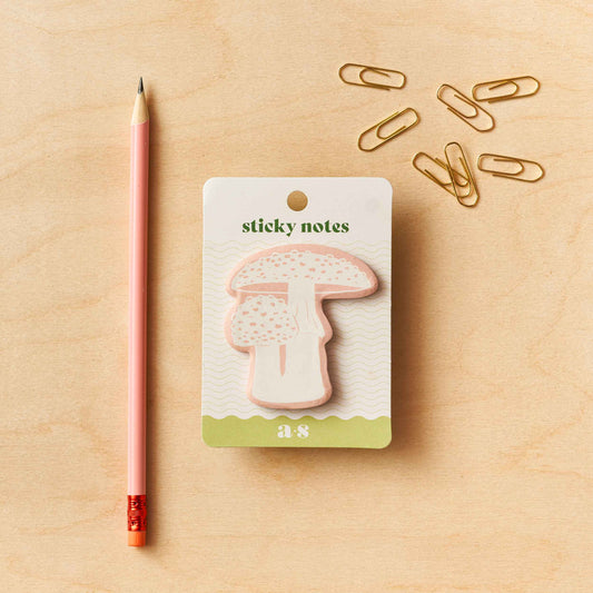 A pack of mushroom-shaped sticky notes by Another Studio with a single pink pen and multiple gold paper clips on a wooden surface.
